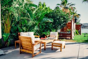 A family arranges patio furniture and cushions in a sunny backyard next to a garden, with trees and a seating area visible.
