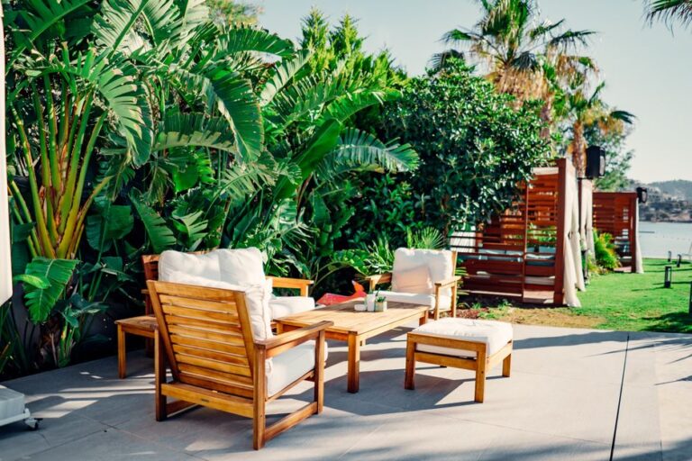 A family arranges patio furniture and cushions in a sunny backyard next to a garden, with trees and a seating area visible.